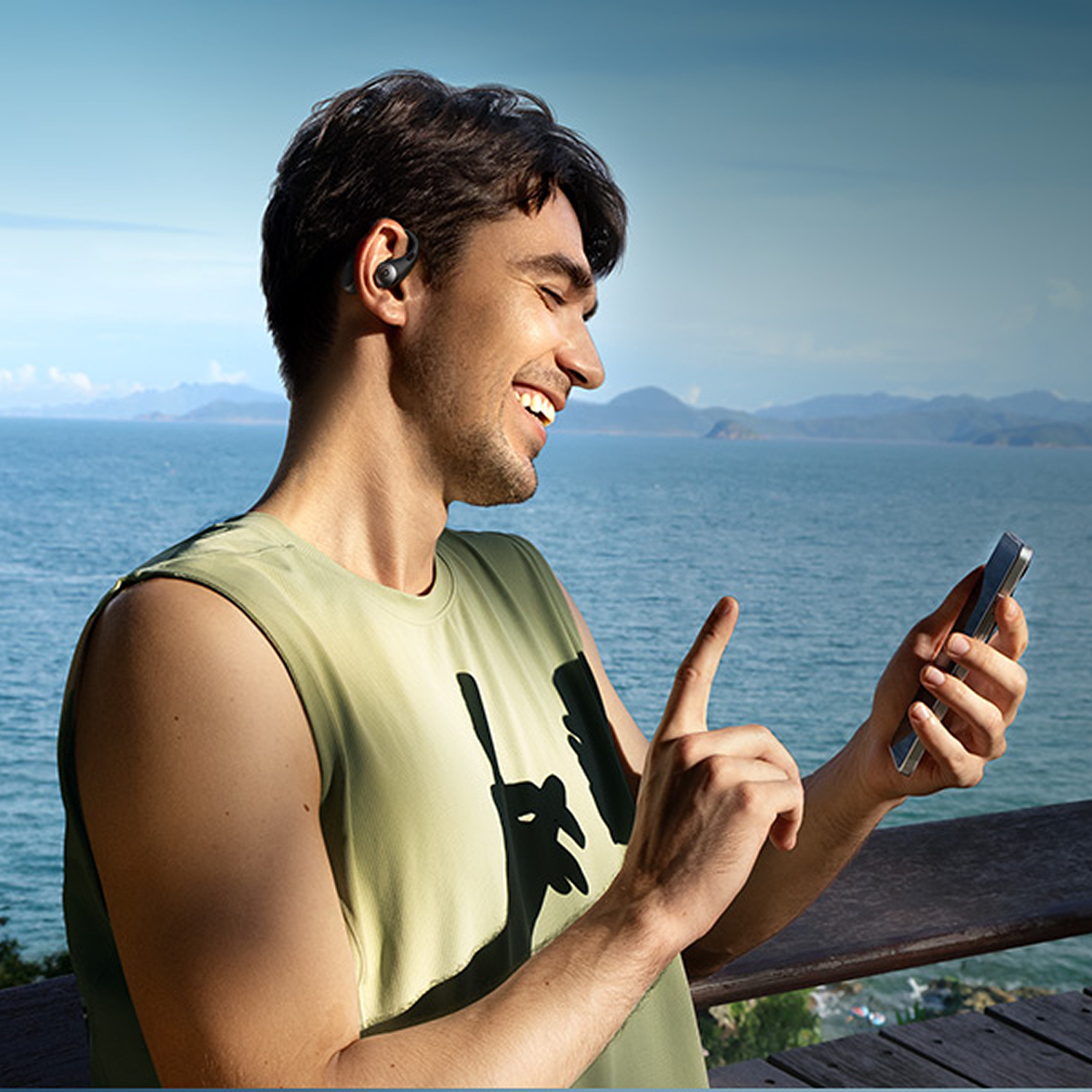 Un hombre sonriente con auriculares sostiene un teléfono y señala junto al mar.
