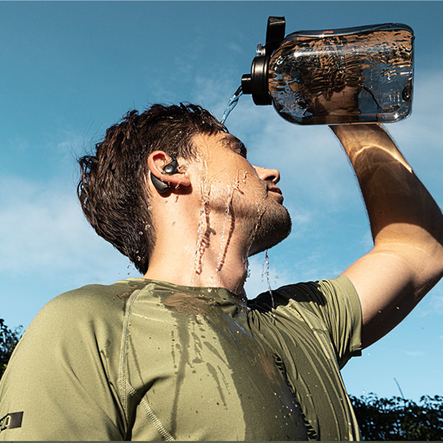 Un hombre se vierte agua de una botella grande en la cara, vistiendo ropa verde al aire libre.