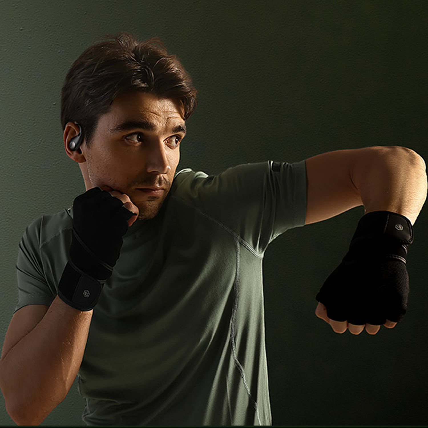 Un hombre con camiseta deportiva verde, auriculares y vendas de boxeo posa en postura de lucha ante una pared oscura.