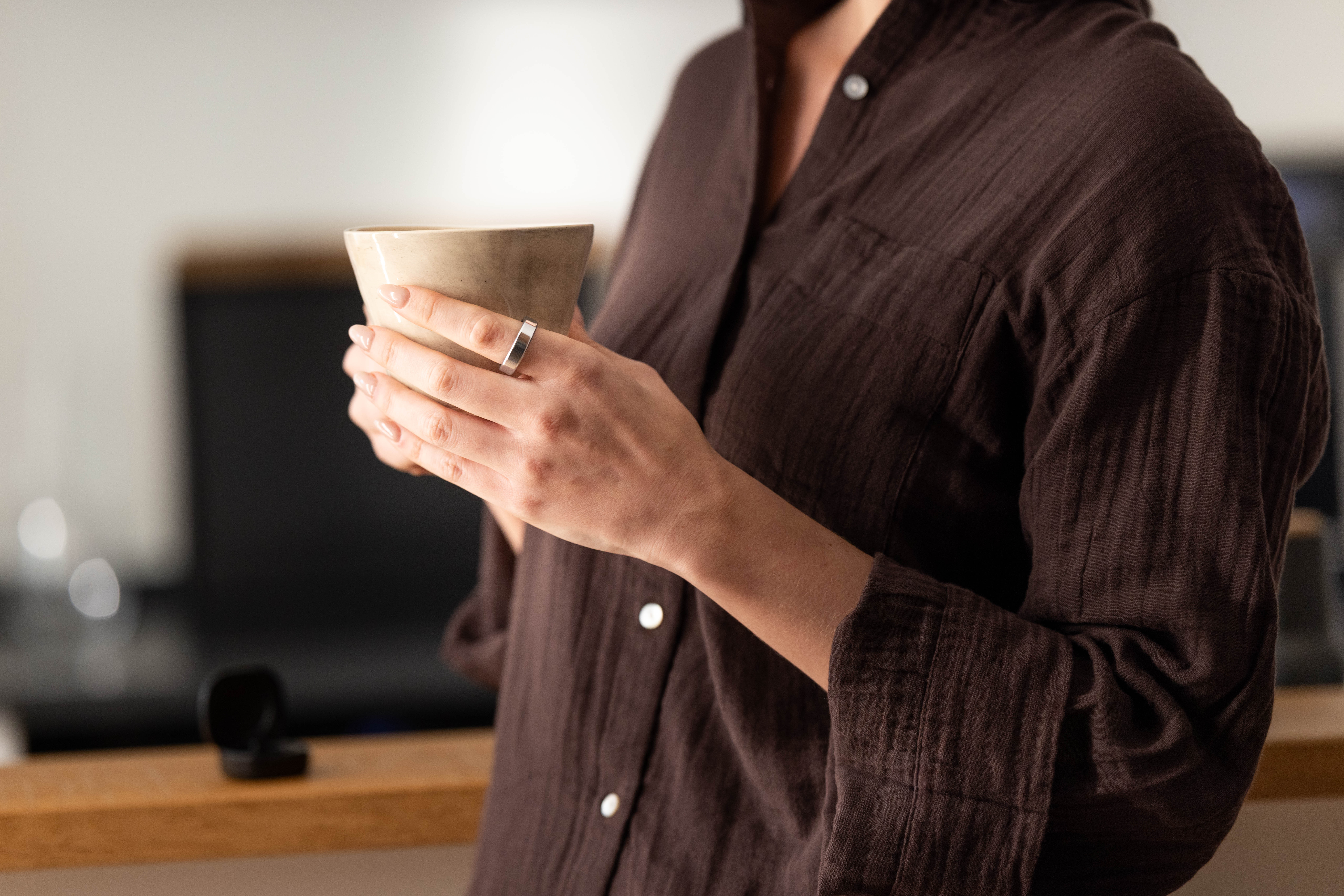 Eine Frau in einem braunen Hemd hält eine Keramiktasse mit beiden Händen, ein glänzender Silberring an ihrem Finger.
