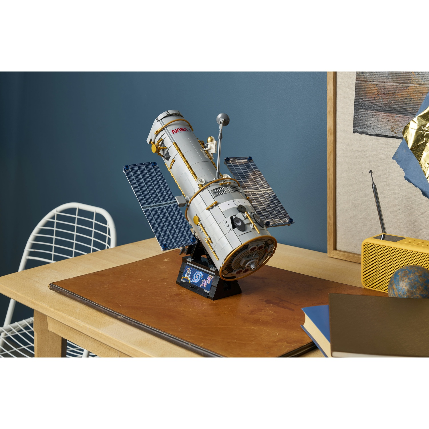 Un dettagliato modello Lego del telescopio spaziale Hubble su una scrivania di legno con libri, una radio e una sedia.