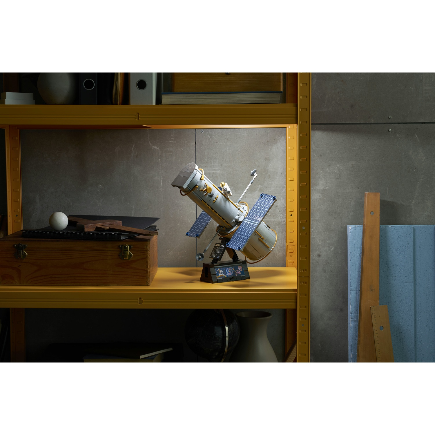 Un modello Lego del telescopio Hubble su uno scaffale giallo contro un muro di cemento grigio.