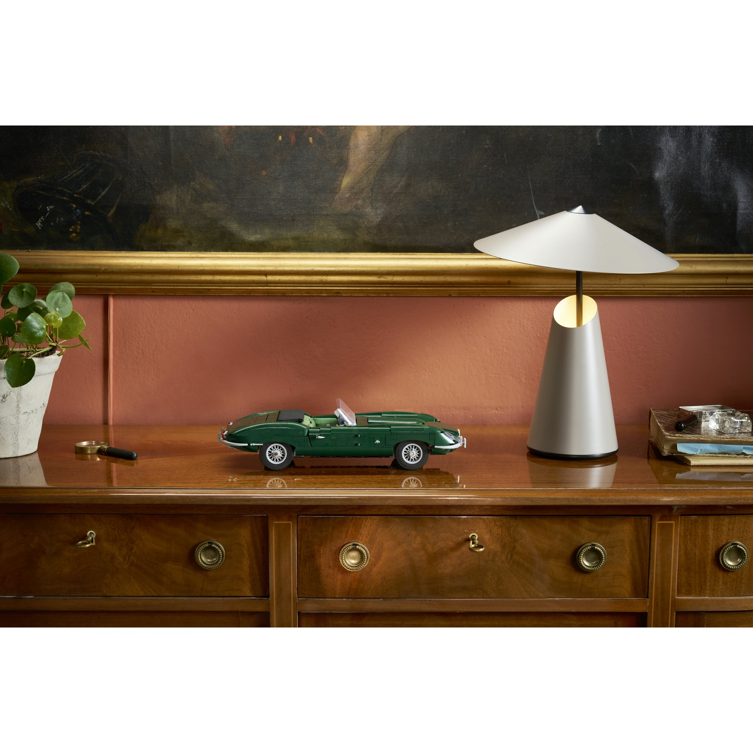 Un'auto giocattolo verde si trova su un comò di legno accanto a una lampada moderna, una pianta e dei libri.