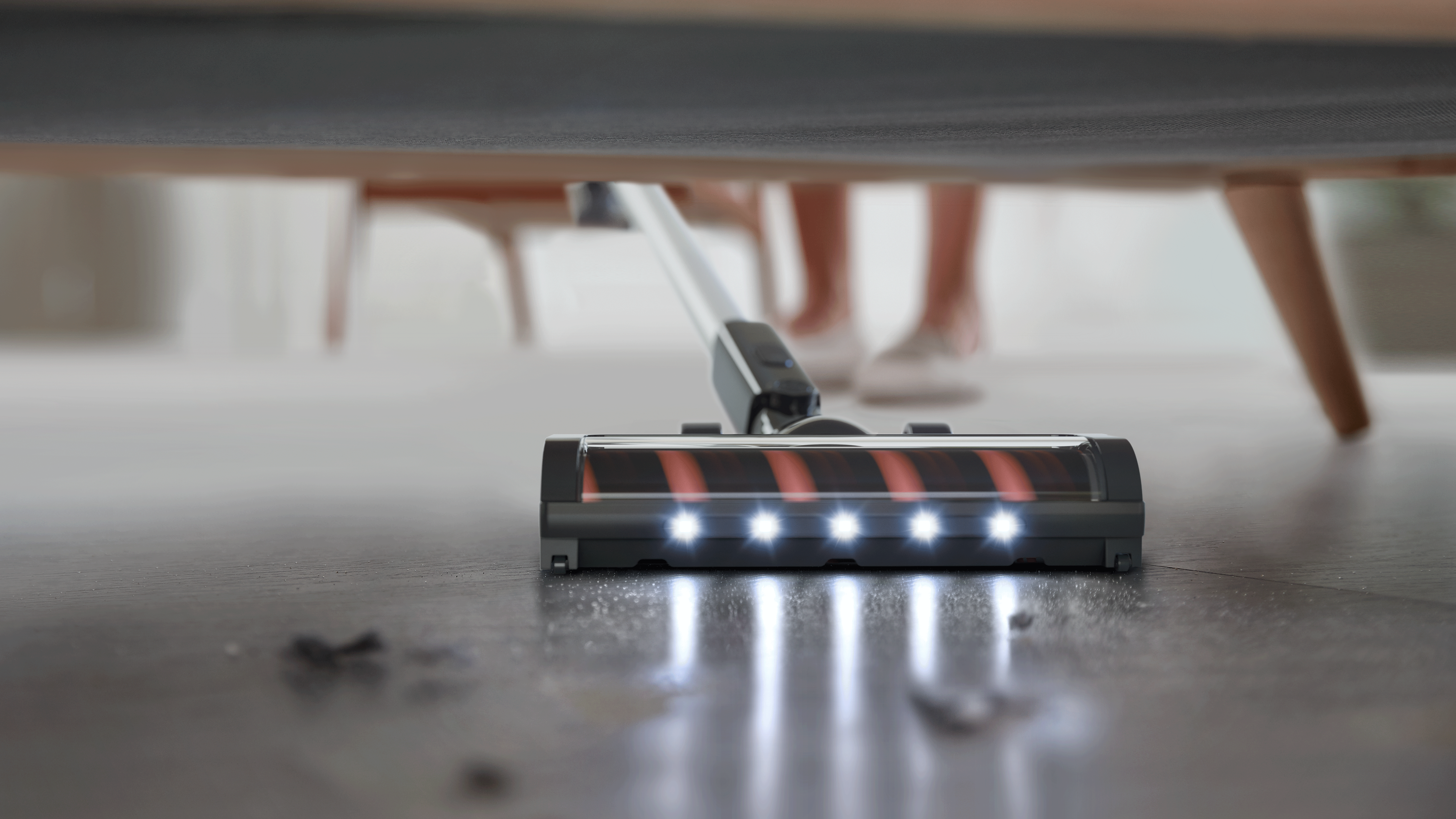 LED-es porszívó takarít a kanapé alatt, a háttérben lábakkal.
