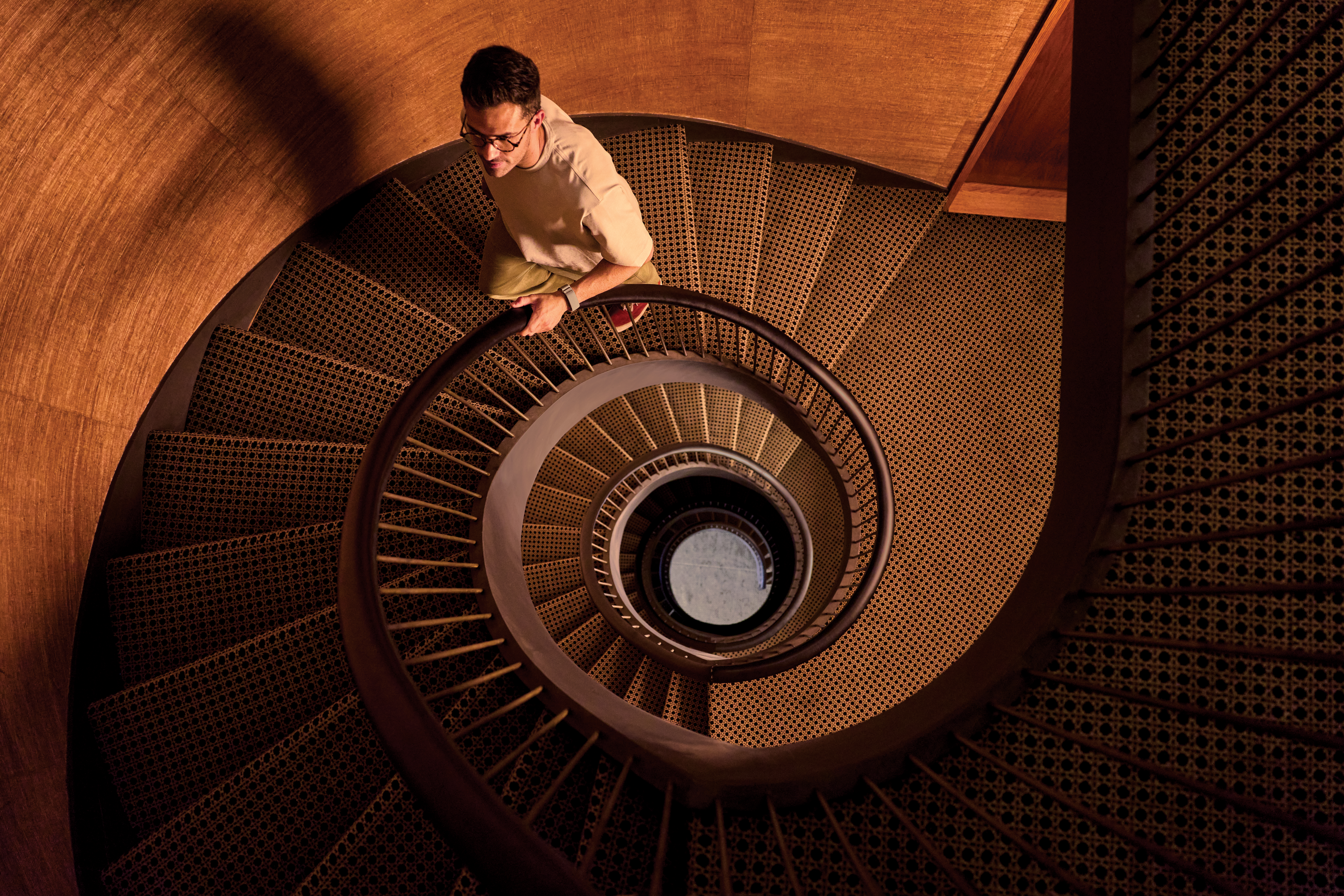 Un homme portant des lunettes monte un élégant escalier en colimaçon lambrissé de bois, vu du dessus.