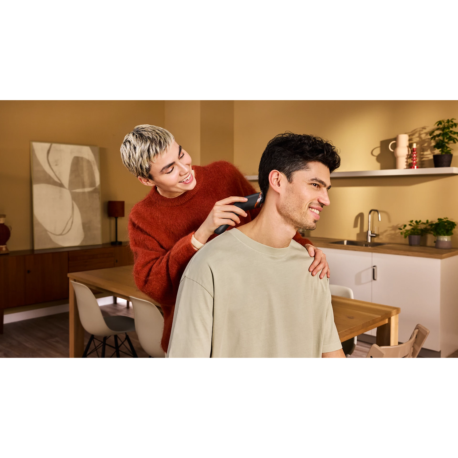 Une personne en pull rouge coupe les cheveux d'un homme avec une tondeuse dans une cuisine.