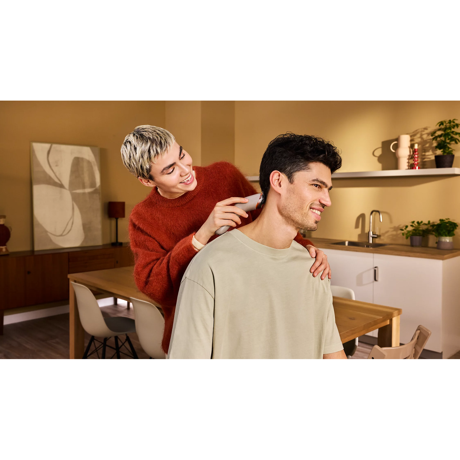 Une personne en pull rouge coupe les cheveux d'un homme à la maison.