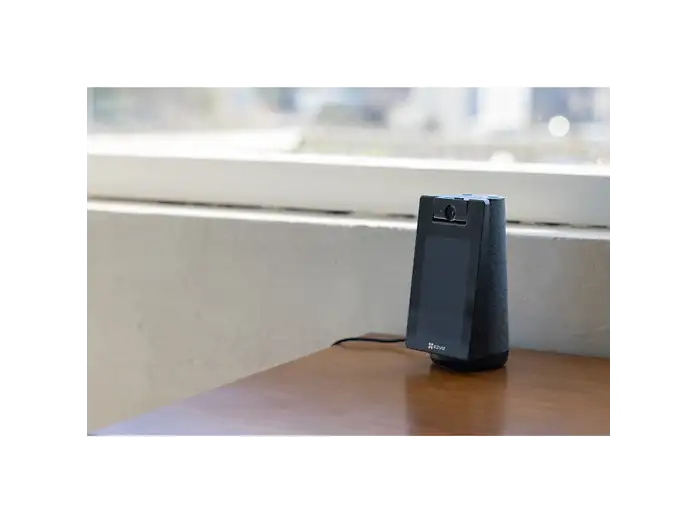 Un dispositivo de seguridad negro con cámara y pantalla sobre una mesa de madera frente a una ventana.