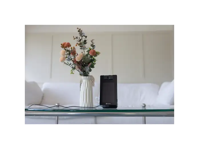 Un dispositivo de seguridad negro y un florero en una mesa de vidrio frente a un sofá blanco.