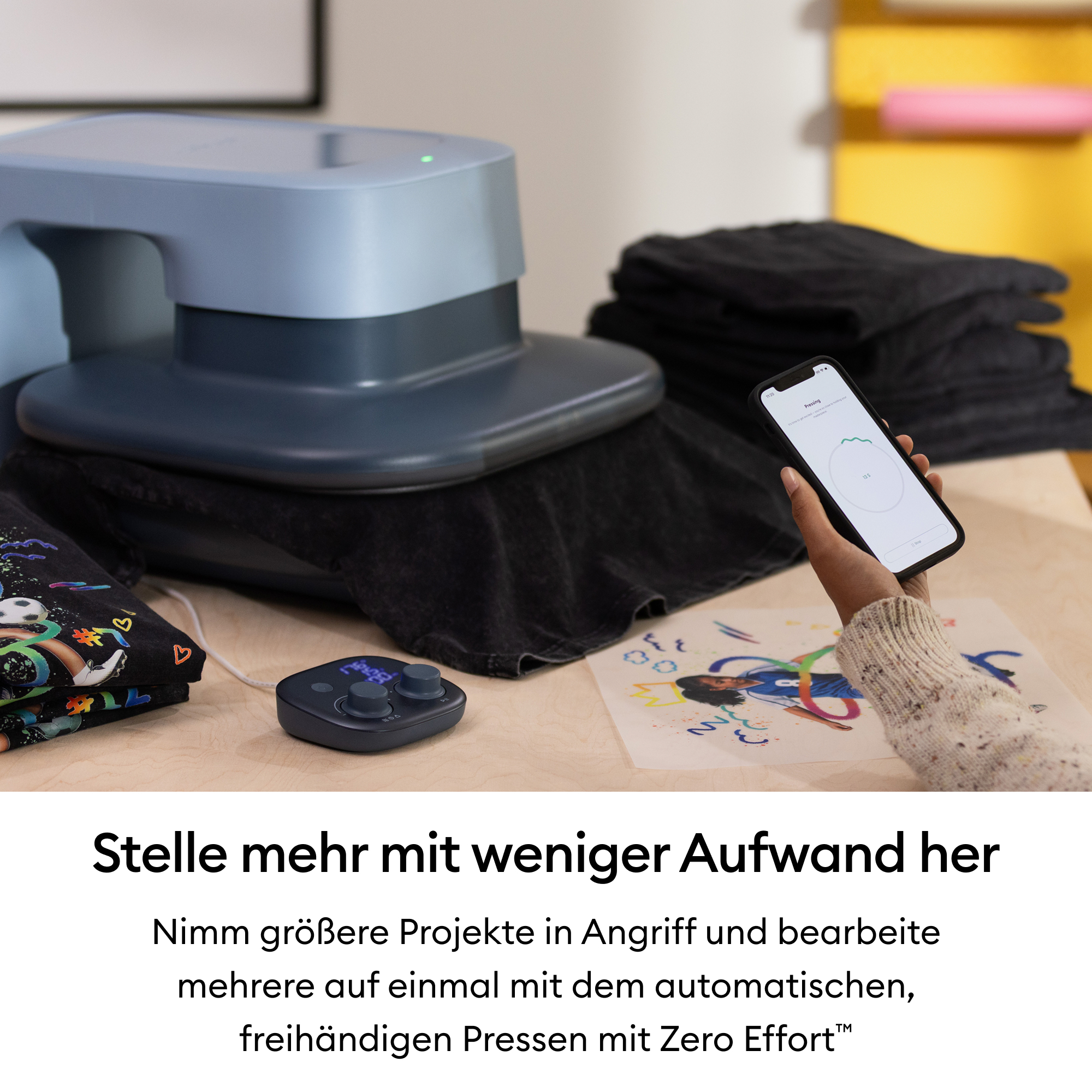 Eine blaue Heißpresse auf einem Tisch mit einem Smartphone, gestapelten schwarzen Hemden und einem Design.