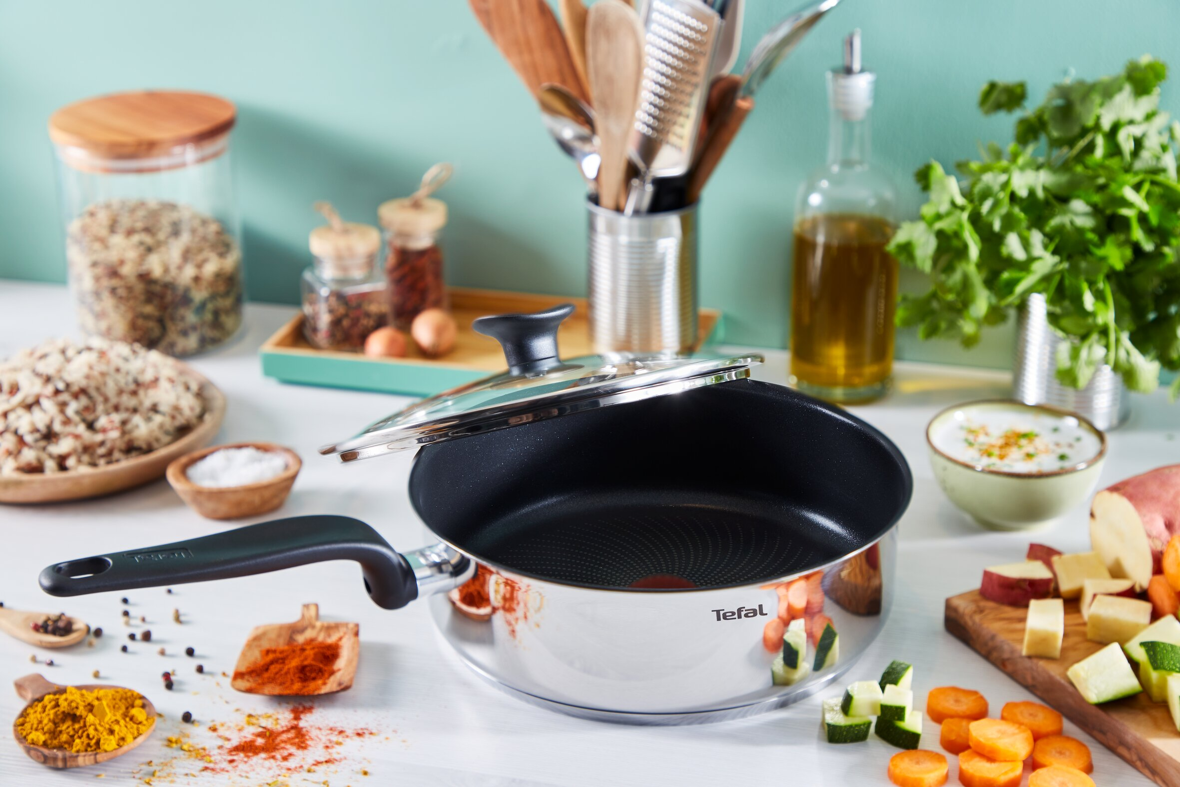 Una sartén de acero inoxidable con tapa de cristal en una encimera de cocina, rodeada de verduras y especias.