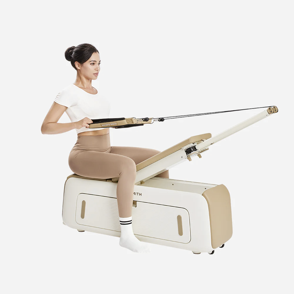 Beyaz tişörtlü ve bej taytlı bir kadın pilates aletine oturmuş direnç iplerini çekiyor.