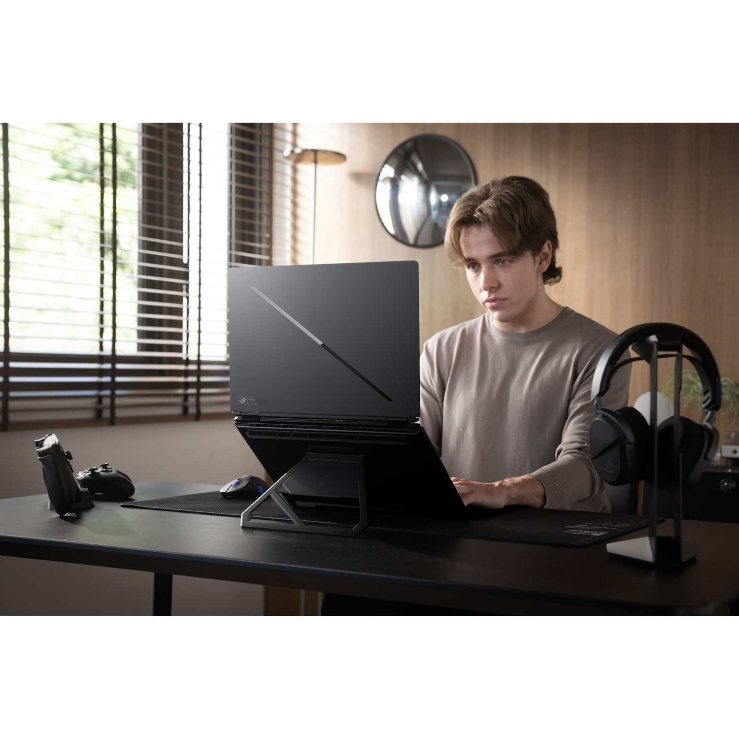 Un giovane siede a una scrivania digitando su un laptop su supporto, con cuffie e controller.