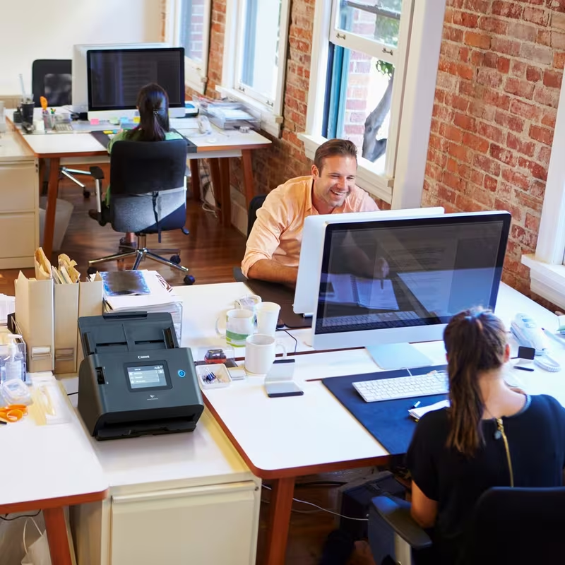 Tre persone lavorano in un ufficio luminoso e moderno con pareti in mattoni e grandi scrivanie bianche. Uno scanner davanti.