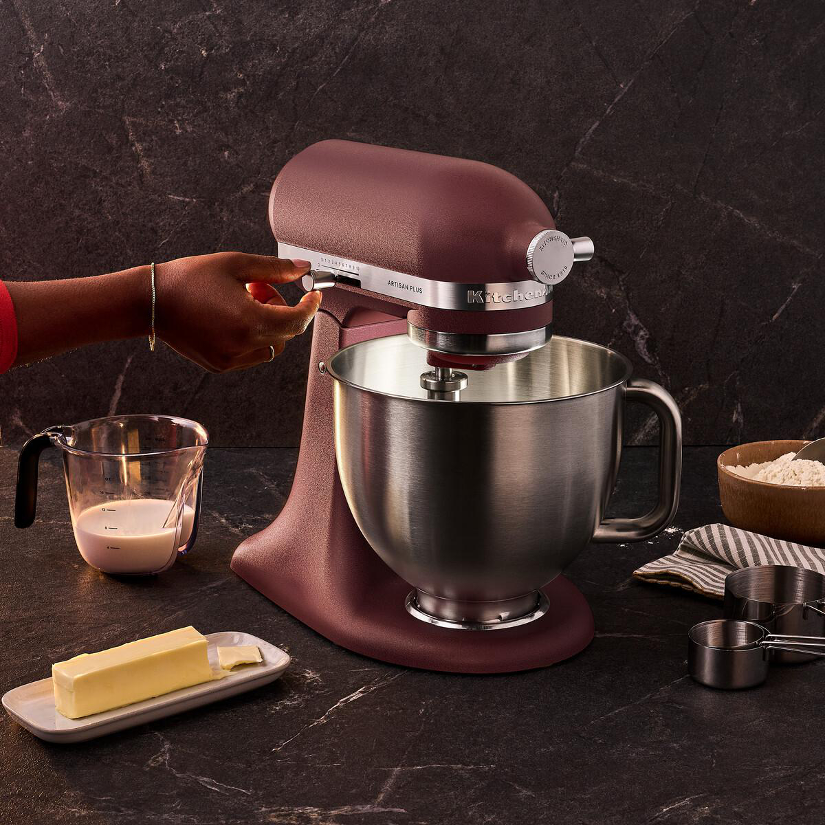 Eine Hand bedient einen weinroten KitchenAid Standmixer auf einer dunklen Steinplatte neben Milch und Butter.