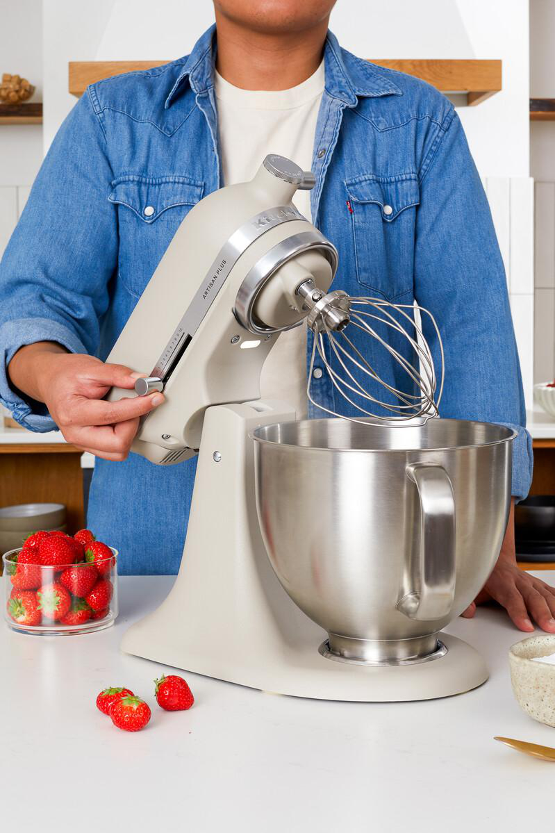 Eine Person im blauen Hemd bedient einen beigen Standmixer auf einer weißen Arbeitsplatte mit frischen Erdbeeren.
