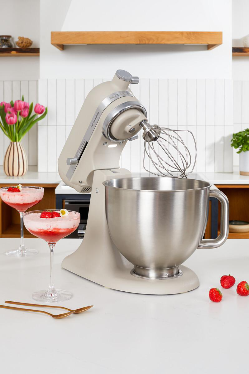 Eine beigefarbene Küchenmaschine auf einer Theke mit zwei Getränken, Erdbeeren und Tulpen im Hintergrund.