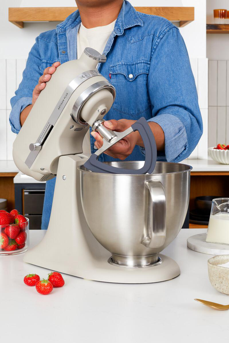 Eine Person befestigt einen Grauen Rührer an einer beigen Küchenmaschine auf einer mit Erdbeeren gedeckten Theke.