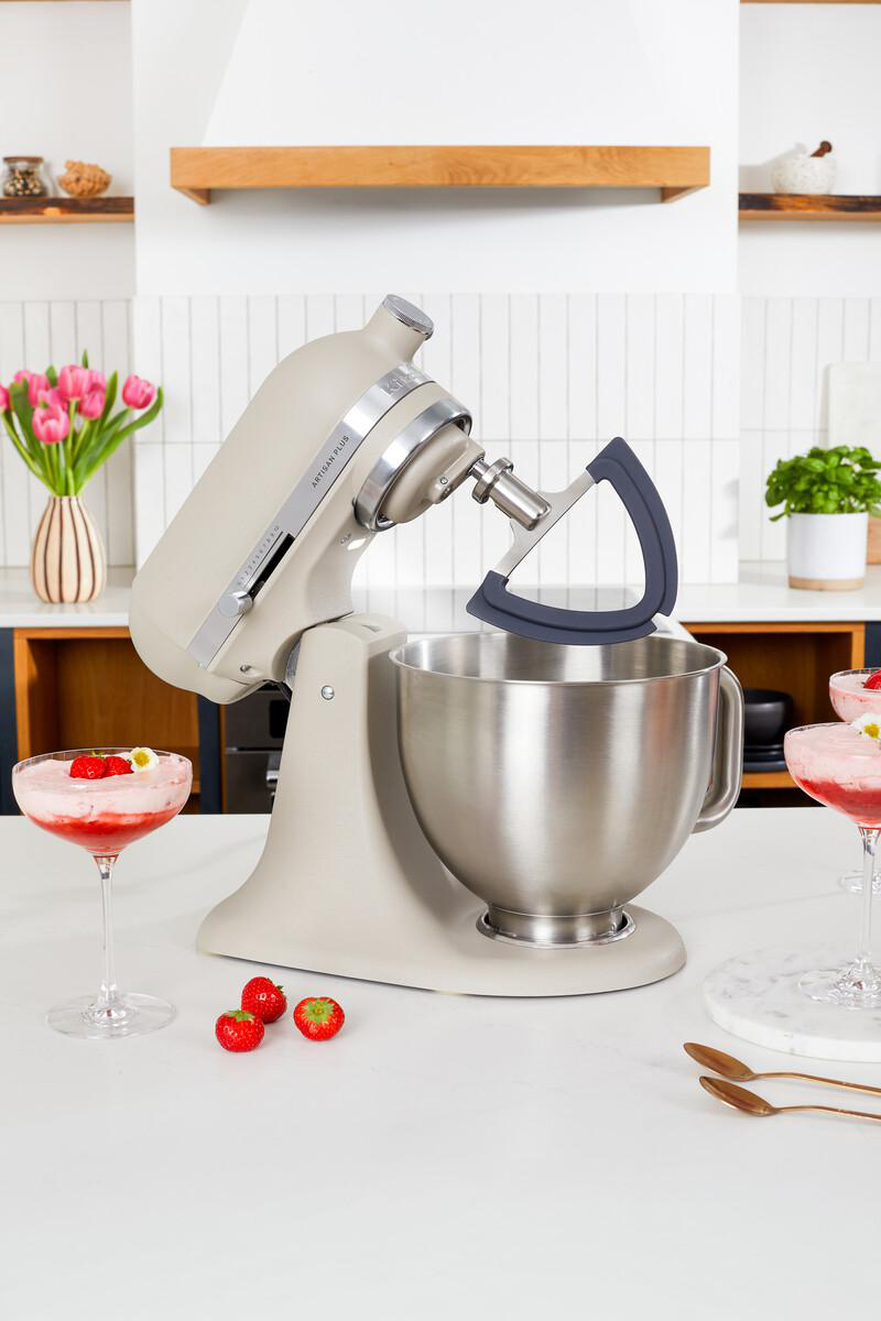 Beigefarbene Küchenmaschine mit Edelstahlschüssel auf einer Theke, umgeben von Erdbeeren und Dessertgläsern.