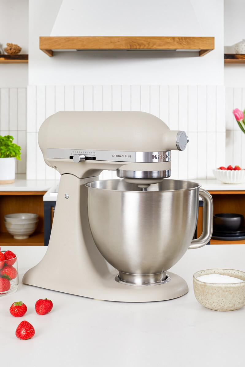 Beigefarbene KitchenAid Artisan Plus Küchenmaschine mit Edelstahl schüssel auf einer Küchenarbeitsplatte, umgeben von Erdbeeren und Zucker.