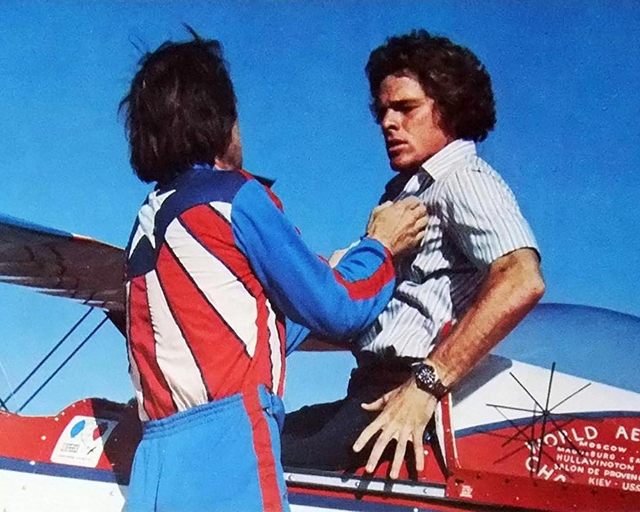Ein Mann im rot-blauen Anzug greift einen anderen Mann am Hemd, der auf einem Flugzeug sitzt.