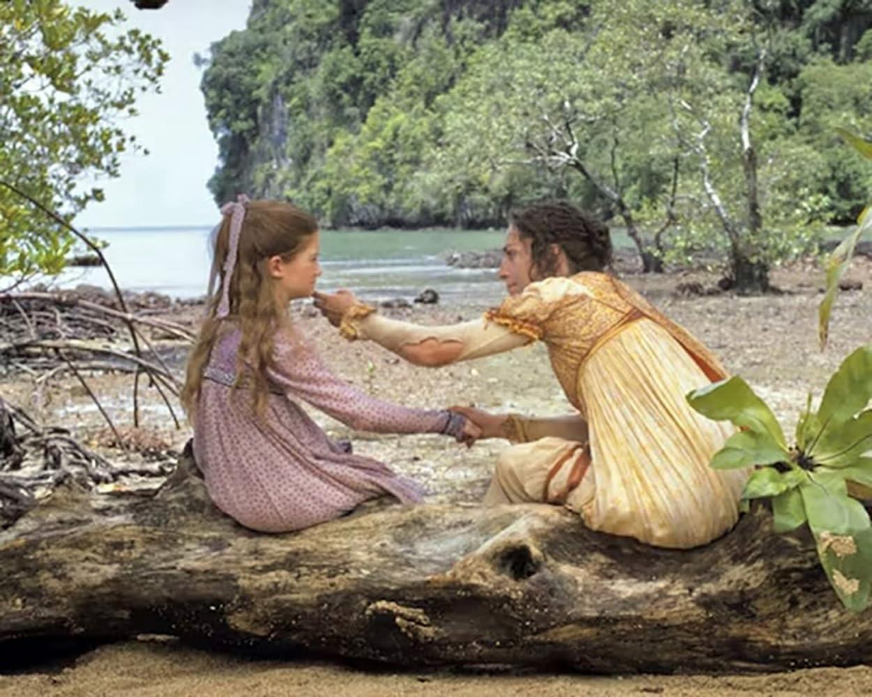 Zwei Frauen in historischen Kleidern sitzen auf einem Baumstamm an einem Strand mit tropischer Vegetation im Hintergrund.
