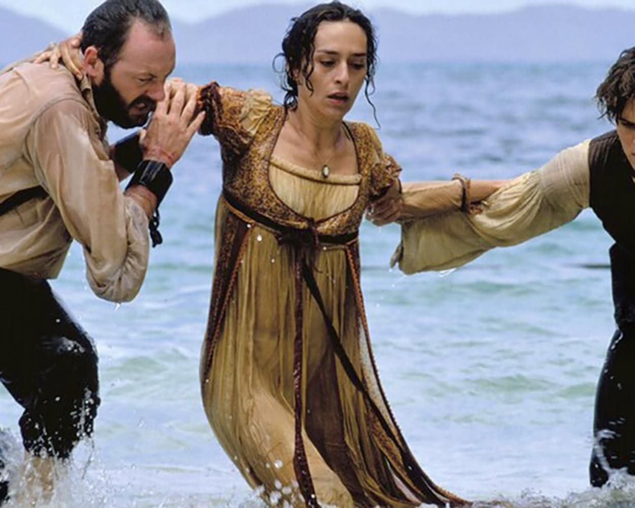 Eine Frau in historischer Kleidung wird von zwei Männern im flachen Wasser am Strand gestützt.