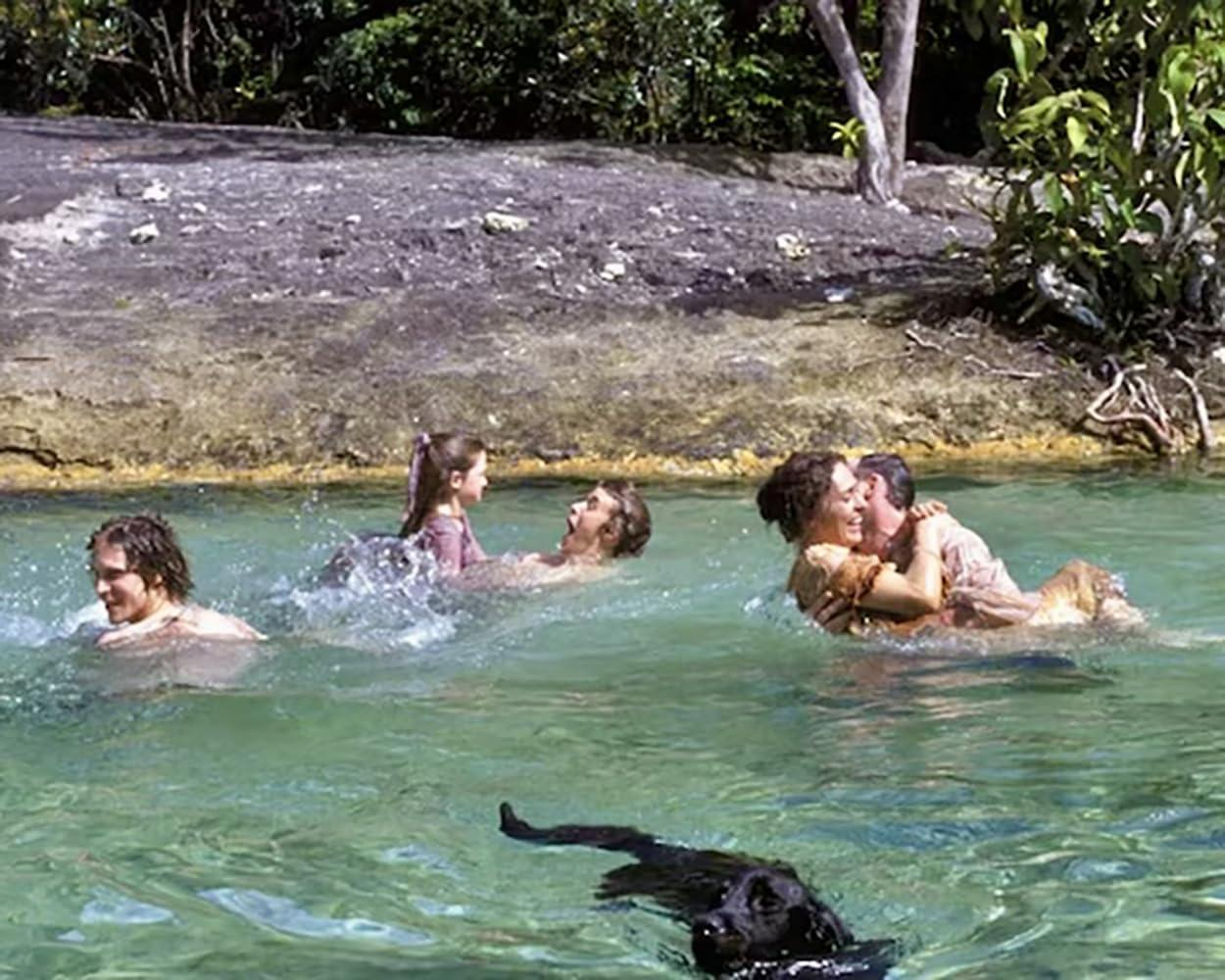 Eine Gruppe von Menschen und ein schwarzer Hund schwimmen im klaren Wasser in der Nähe eines felsigen Ufers.