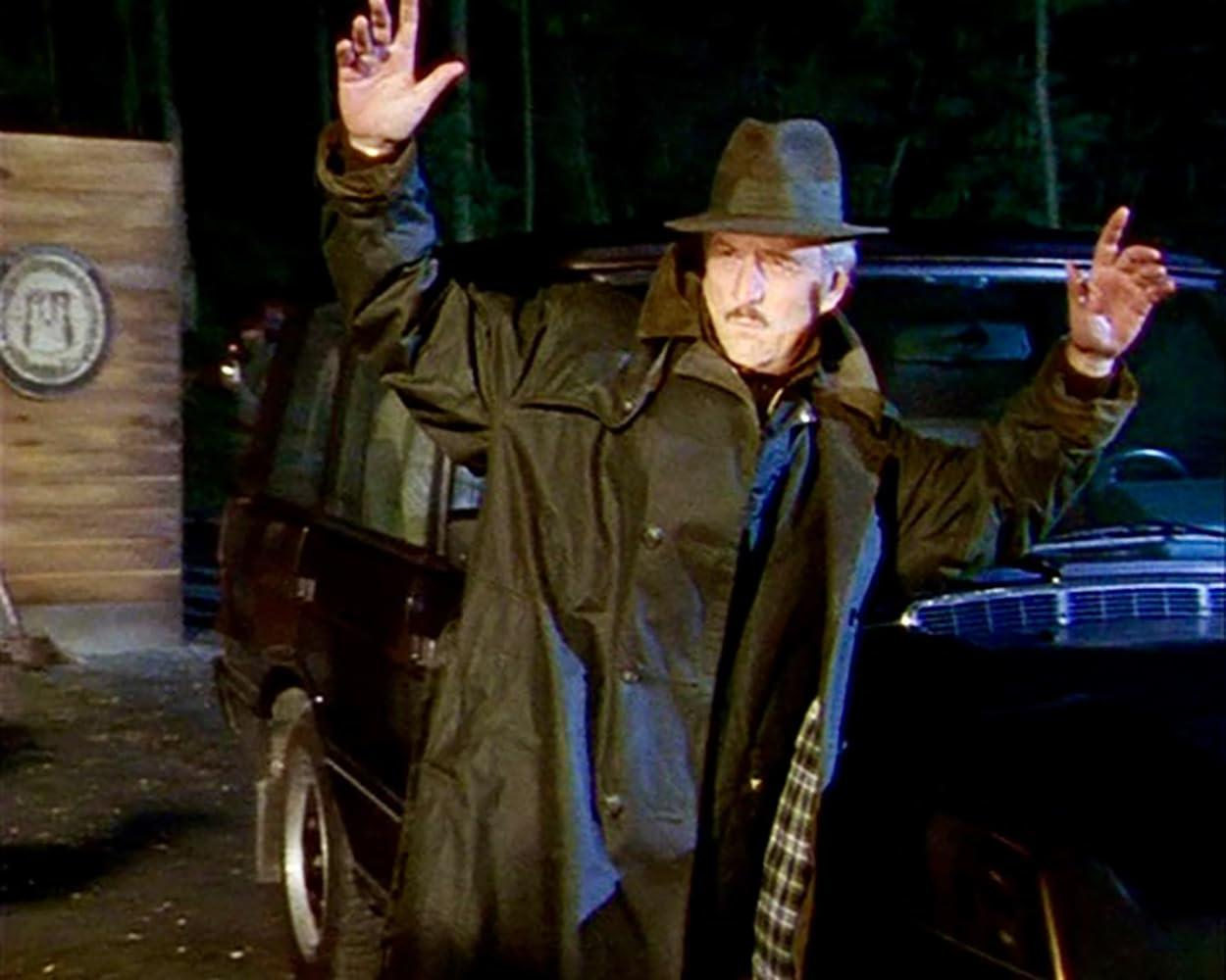 Ein Mann im dunklen Trenchcoat und Hut steht nachts mit erhobenen Händen an einem schwarzen Auto.