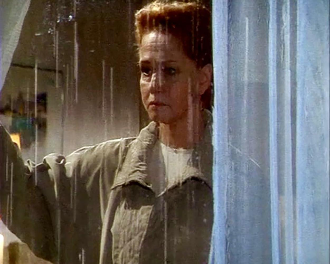 Eine Frau mit kurzen roten Haaren und Jacke steht hinter einer regennassen Fensterscheibe neben einem blauen Vorhang.