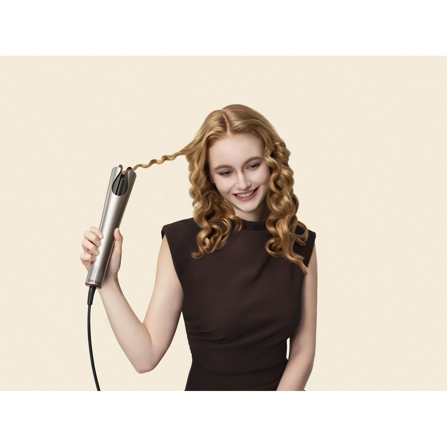 Une femme blonde souriante utilise un fer à friser automatique argenté sur une mèche de cheveux.