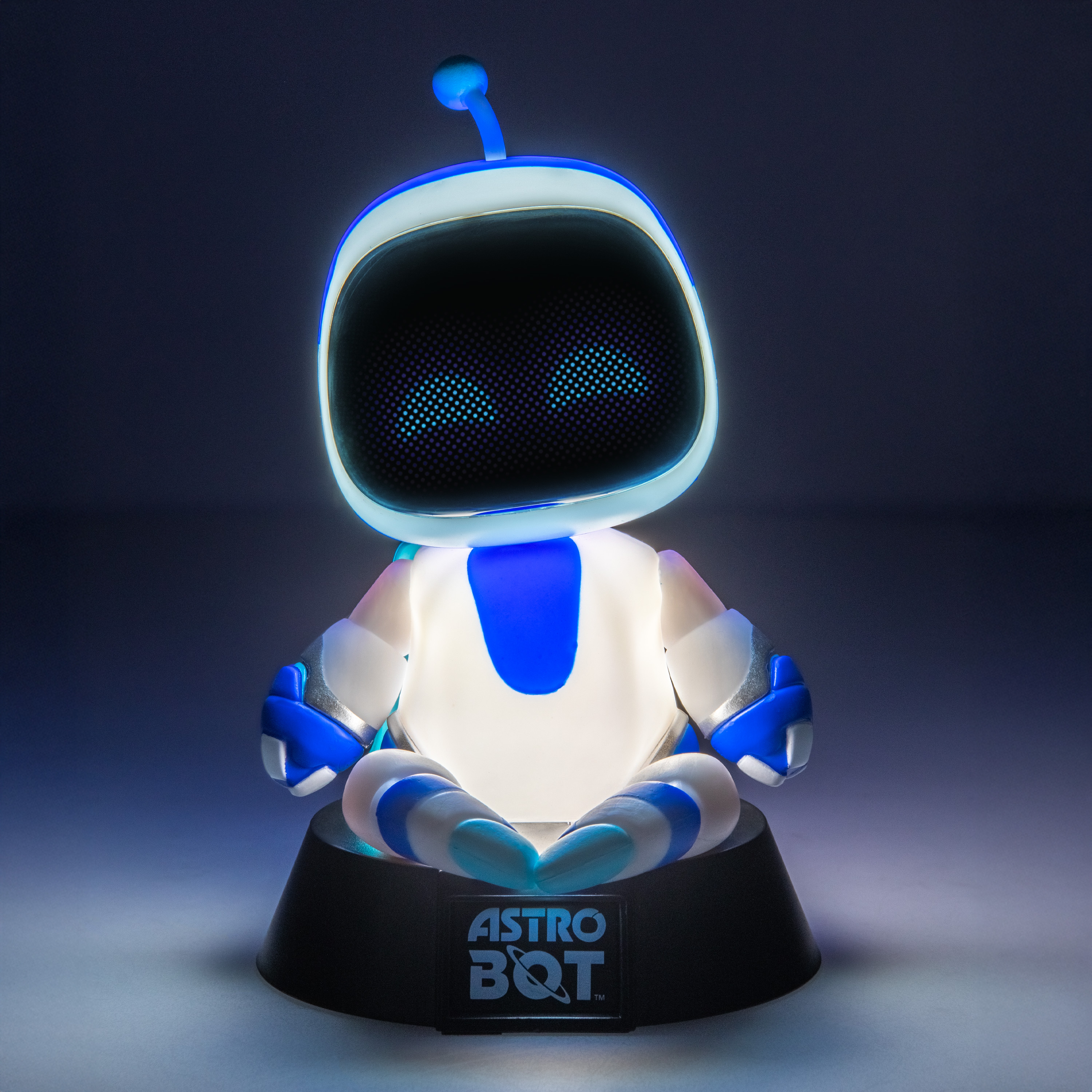 Eine leuchtende Astro Bot-Figur sitzt im Schneidersitz auf einem schwarzen Ständer vor dunklem Hintergrund.