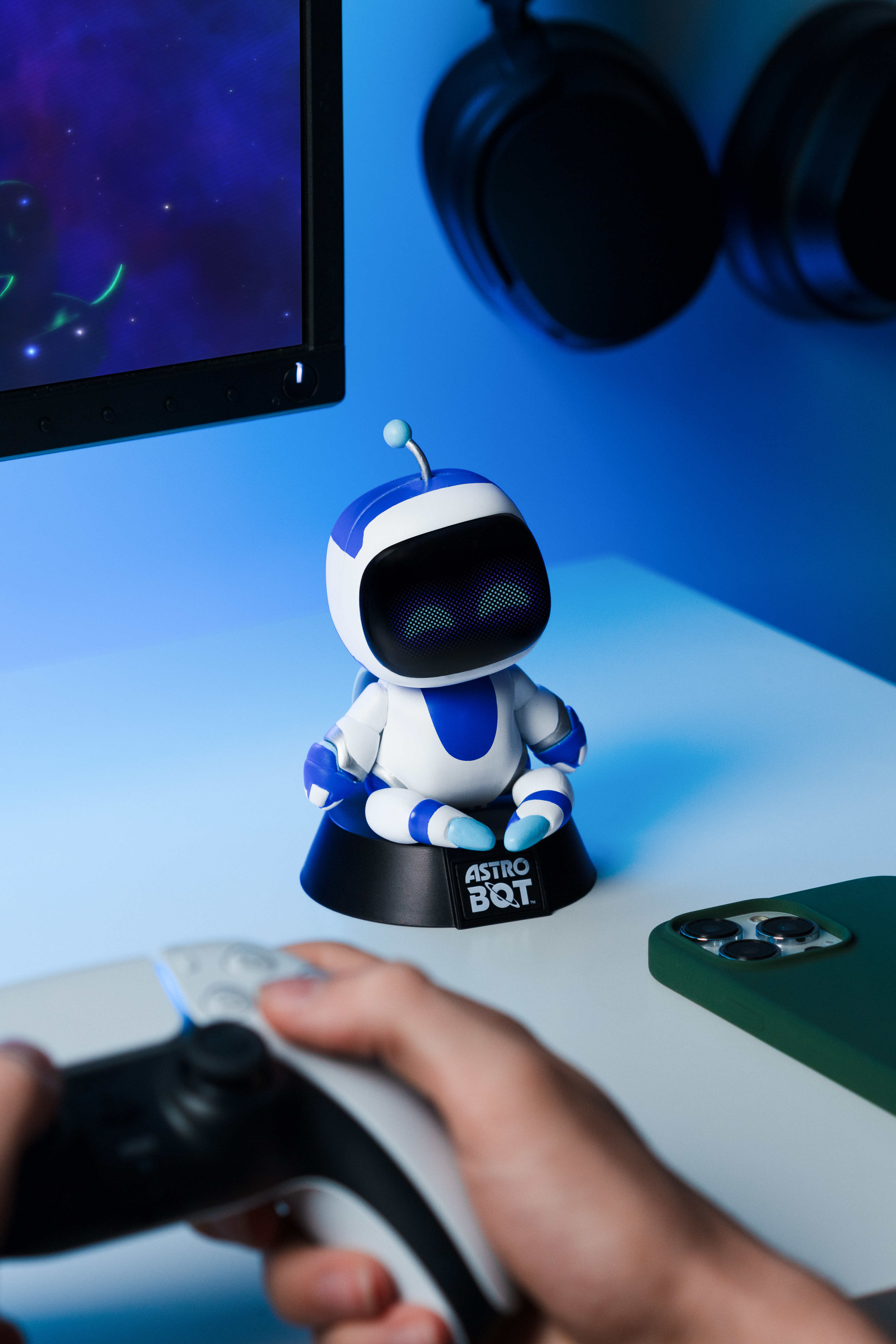Eine Astro Bot Figur sitzt auf einem Stand vor einem Bildschirm, während jemand einen Controller hält.