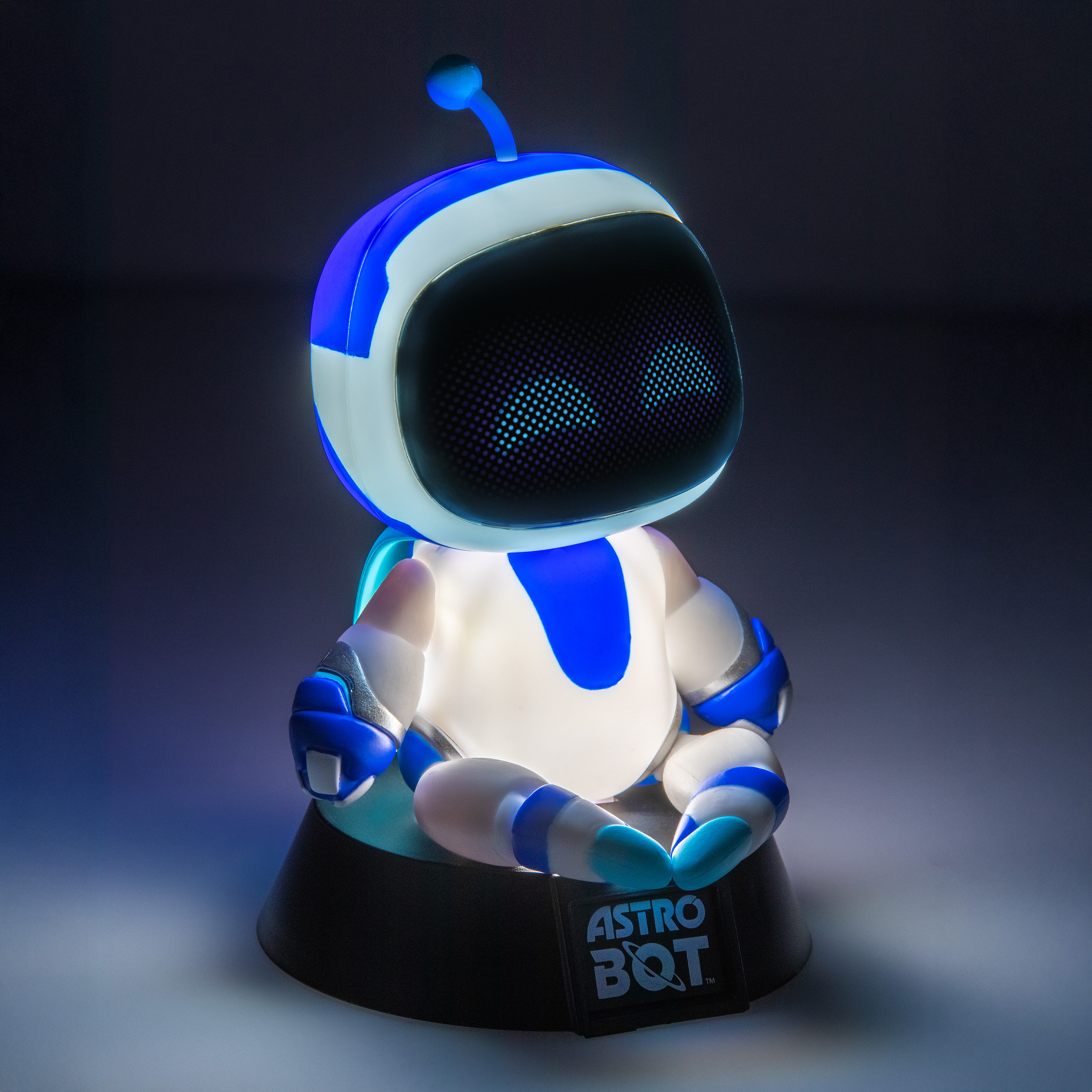 Beleuchtete Astro Bot Figur in blau und weiß sitzt im Schneidersitz auf einem dunklen Sockel vor grauem Hintergrund.