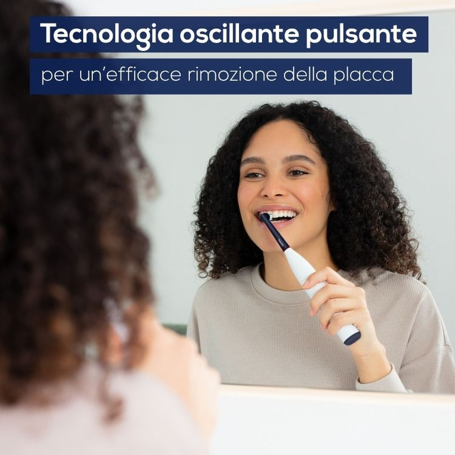 Una donna con i capelli scuri e ricci si guarda allo specchio mentre si lava i denti con uno spazzolino elettrico.