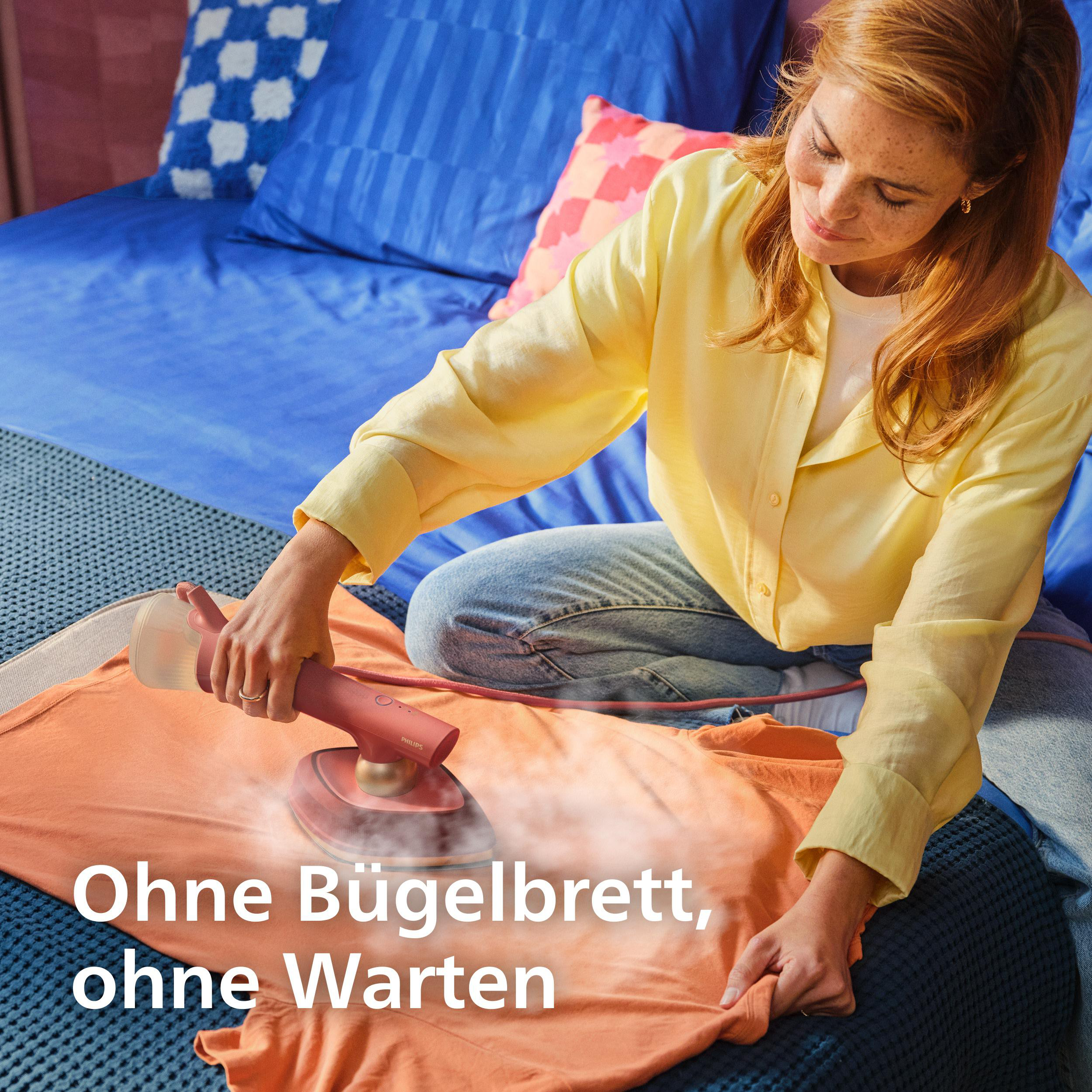 Eine Frau bügelt ein orangefarbenes Kleidungsstück auf dem Bett mit einem Dampfglätter, Text: Ohne Bügelbrett, ohne Warten
