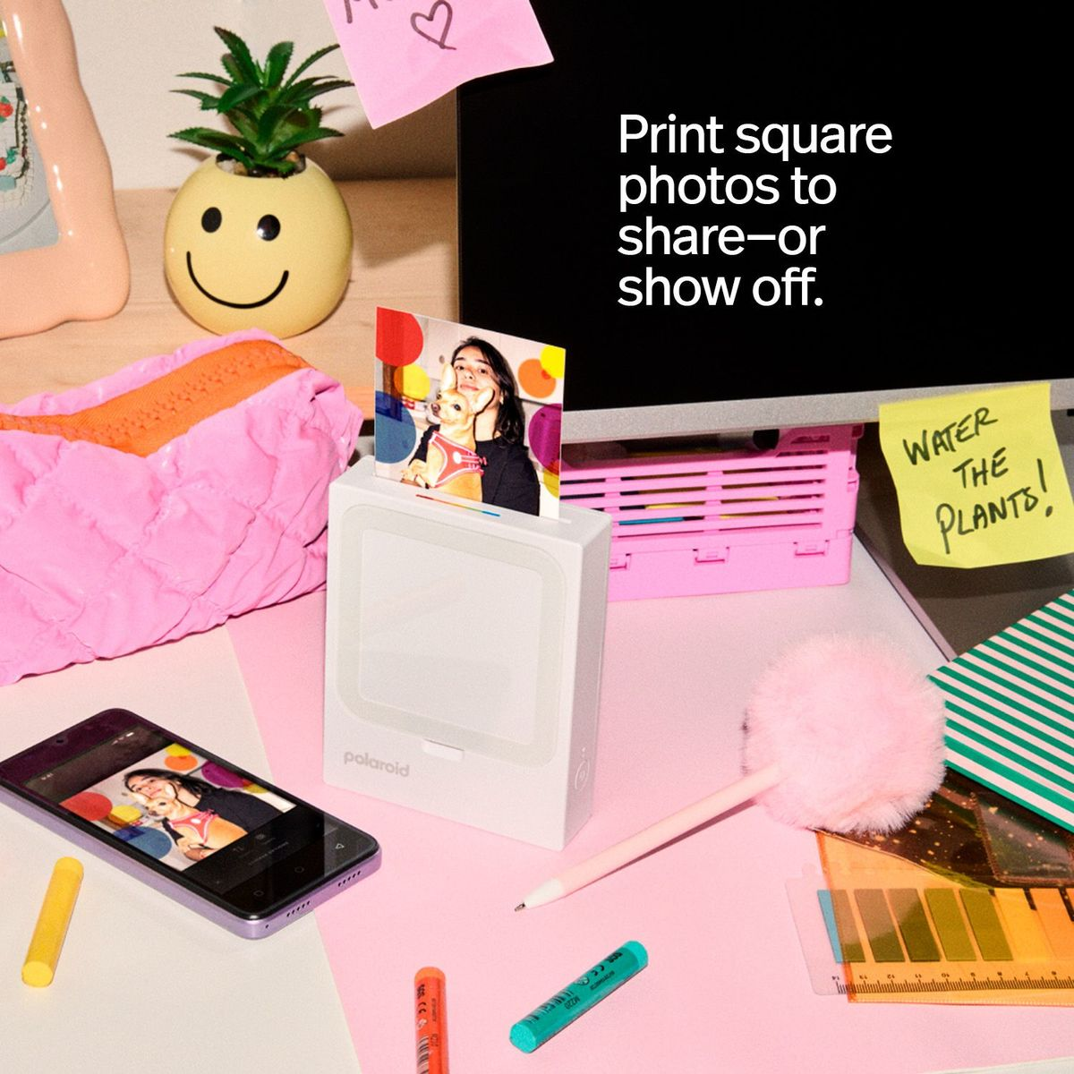 Ein Polaroid-Drucker auf einem Schreibtisch mit druckendem Foto, einem Smartphone, bunten Stiften, einem Smiley-Blumentopf.