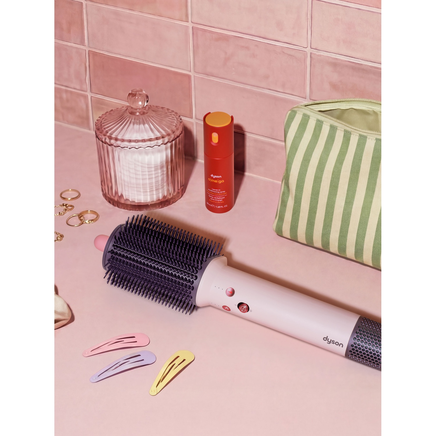 Uno styler Dyson su superficie rosa con mollette, un barattolo, uno spray e una borsa a righe.