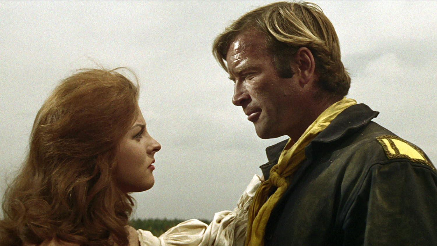 Ein blondhaariger Mann und eine rothaarige Frau stehen nah beieinander vor einem hellen Himmel.