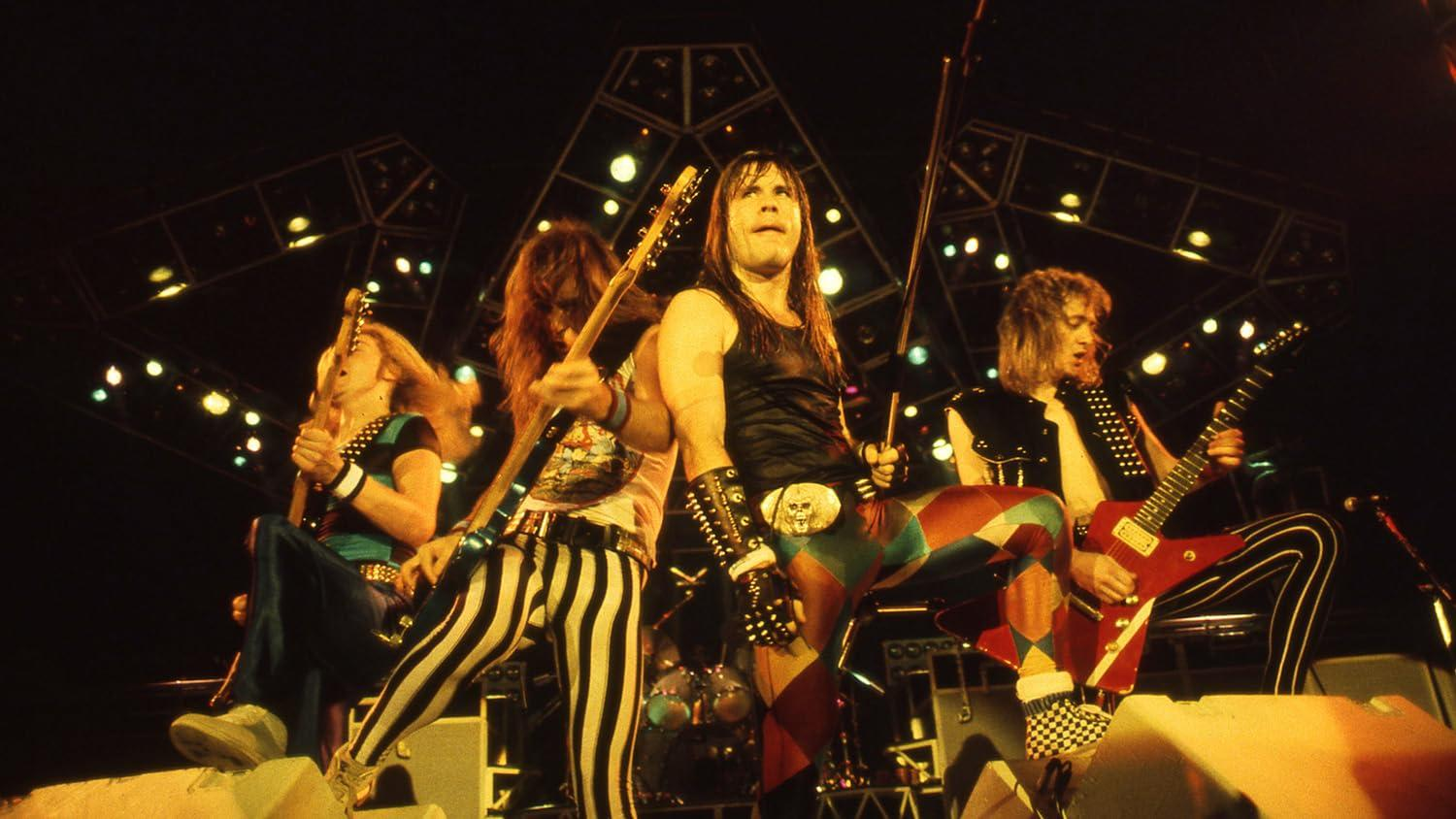 Vier langhaarige Rockmusiker mit Gitarren auf einer hell erleuchteten Bühne.