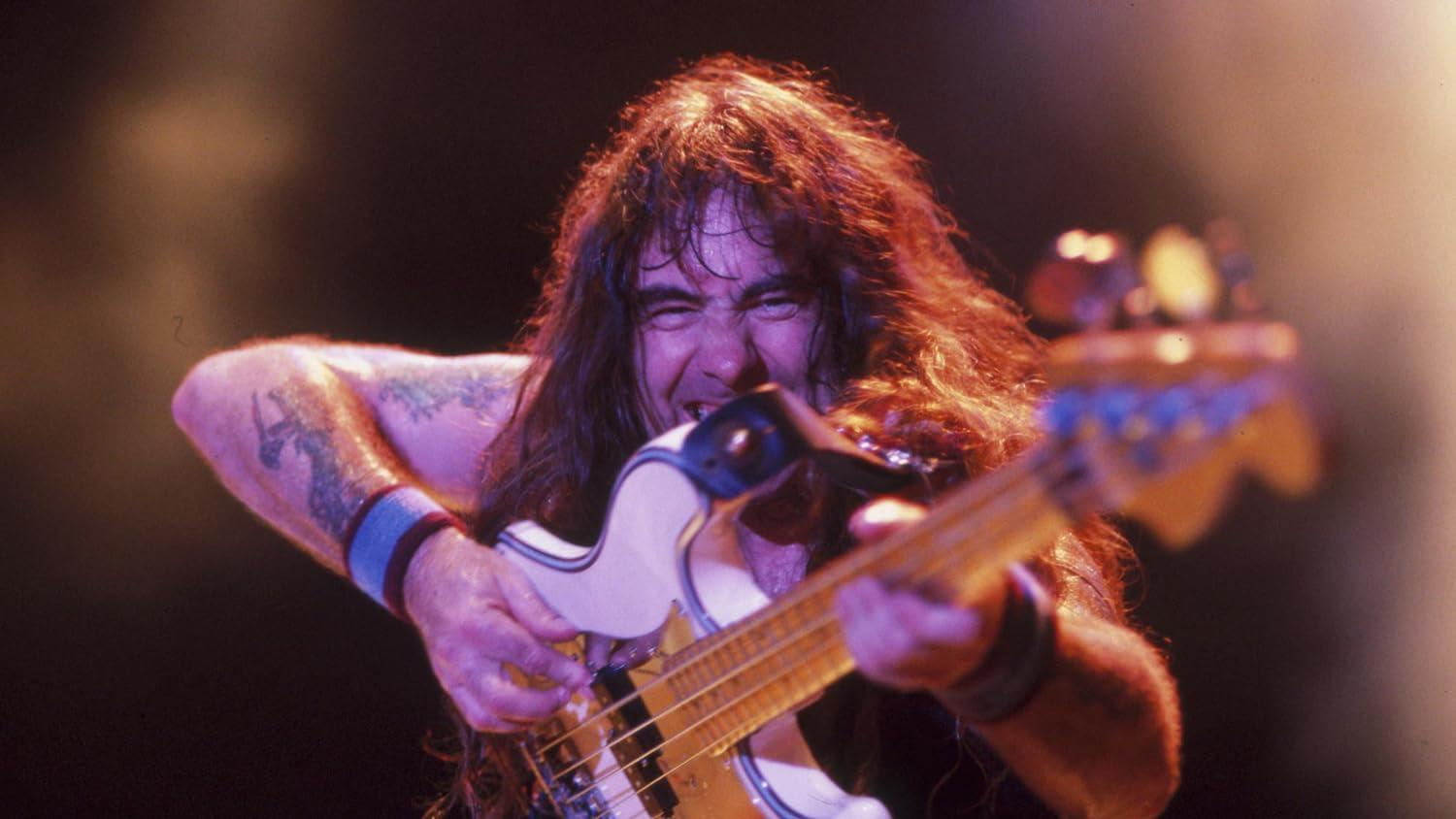 Ein langhaariger Bassist spielt energisch auf einer weißen E-Bassgitarre unter Bühnenbeleuchtung.