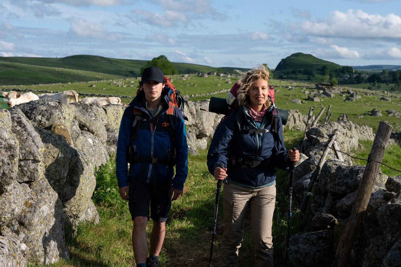 Zwei Wanderer mit Rucksäcken gehen auf einem Weg zwischen alten Steinmauern in einer hügeligen Landschaft.