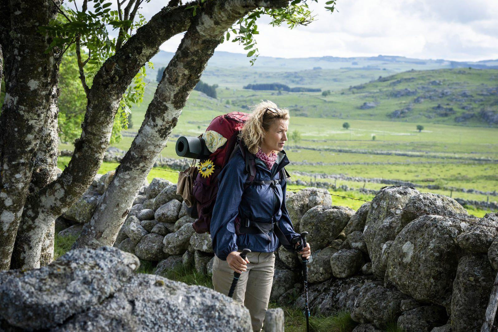 Wanderin mit großem Rucksack und Stöcken geht an einer Steinmauer in hügeliger Landschaft vorbei.