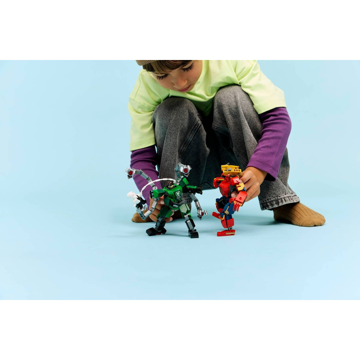 Un bambino in jeans e maglietta verde gioca con due action figure Lego su sfondo azzurro.