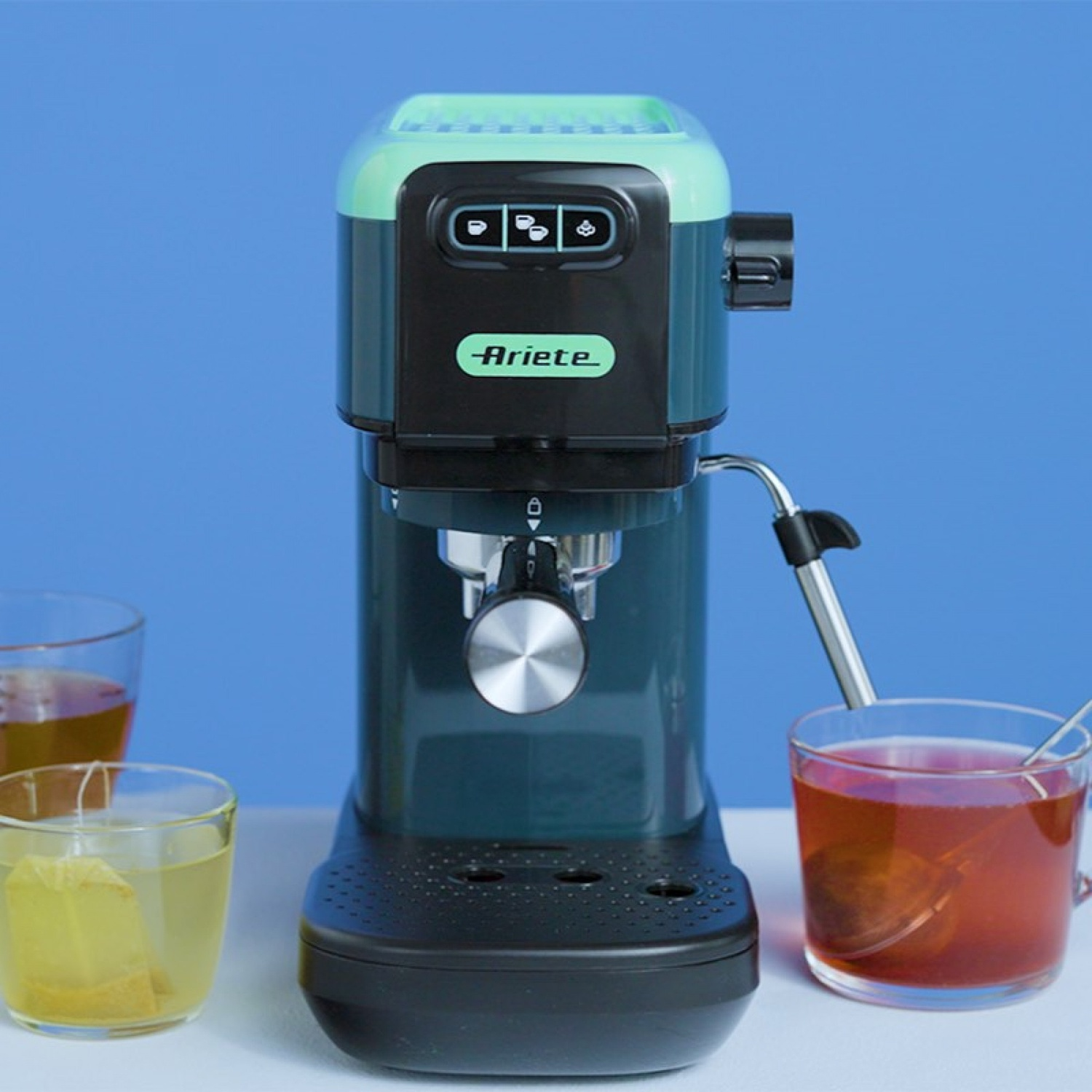 Una macchina da caffè espresso Ariete verde su un tavolo bianco con sfondo blu, affiancata da bicchieri di tè.