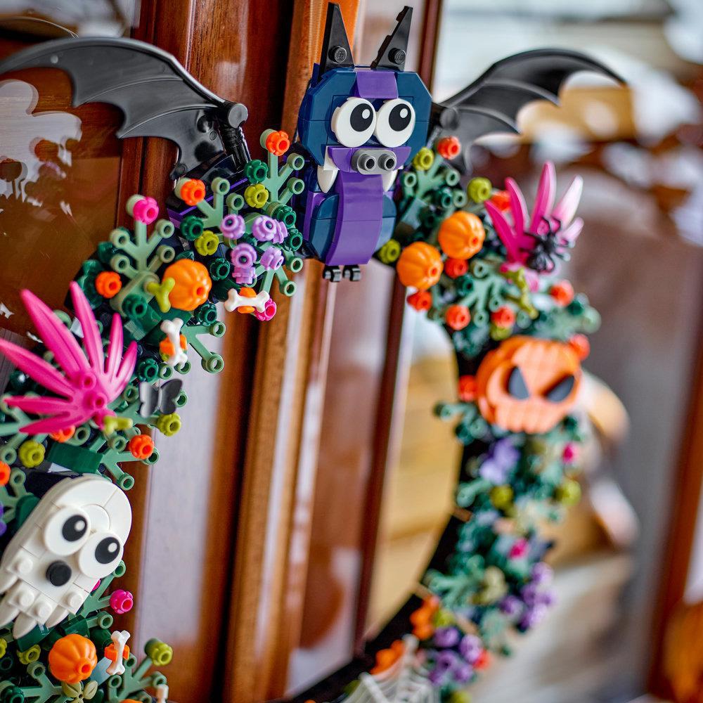 Ein Lego-Halloween-Kranz an einer Holztür zeigt eine violette Fledermaus oben, einen weißen Geist, Kürbisse und rosa Blätter.