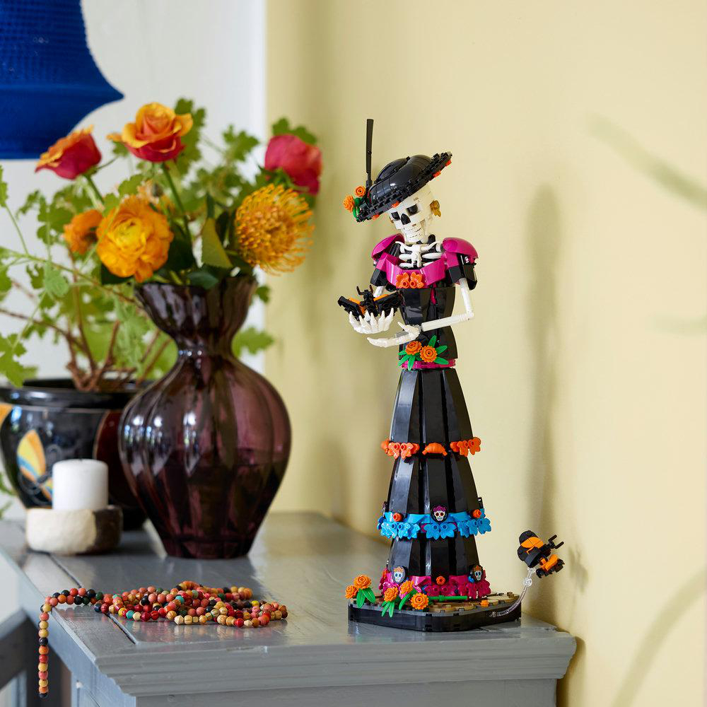 Eine Lego-Skelettfigur im eleganten Kleid steht auf einem grauen Tisch neben einer Vase mit bunten Blumen.