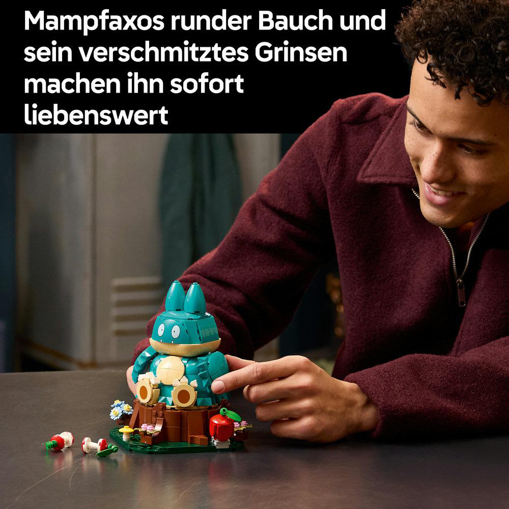 Ein lächelnder Mann baut eine blaue LEGO Snorlax Figur auf einem verzierten Baumstumpf mit deutschem Text.