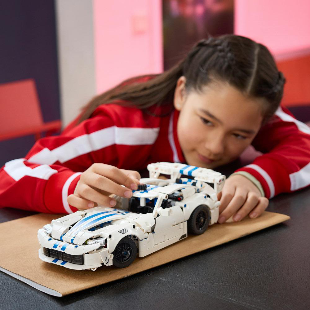 Ein junges Mädchen in roter Jacke baut ein weiß-blaues Lego-Modellauto auf einem Tisch zusammen.