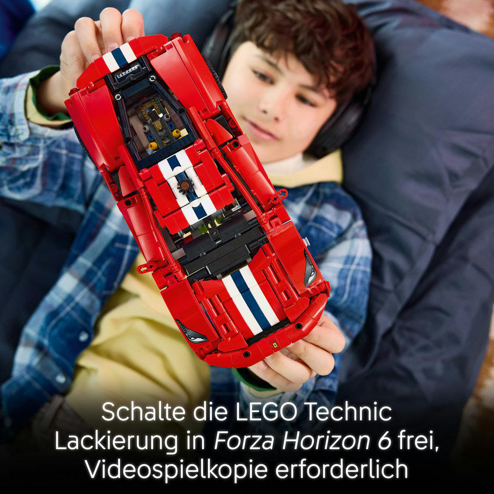 Ein Junge hält ein rotes Lego Technic Auto mit weißen Streifen vor sich, während er auf einem Kissen sitzt.
